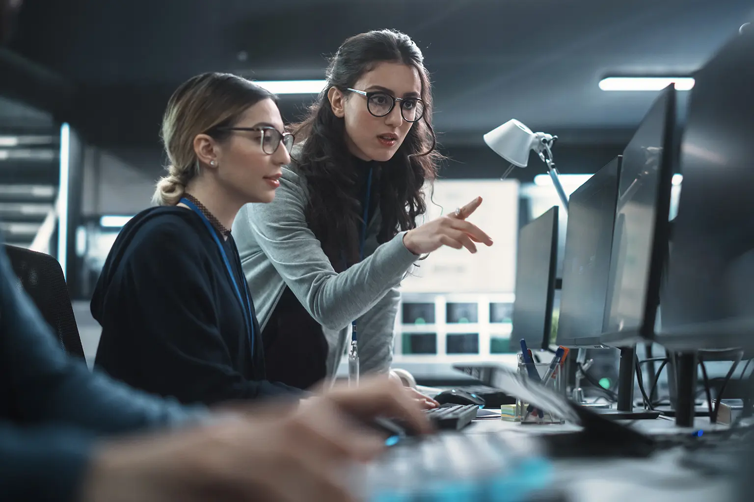 Two women at a computer workstation, one seated and the other pointing at a monitor, working together in a dimly lit office environment.