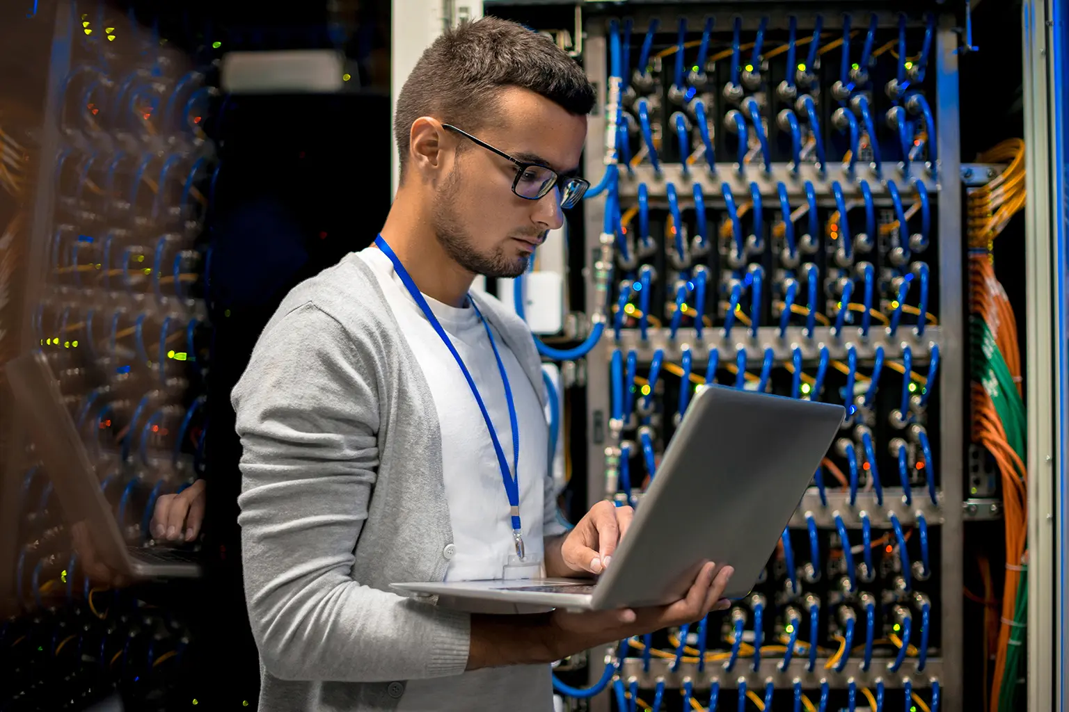 A man with glasses works on a laptop in a server room, surrounded by cables and equipment.
